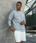 A man wearing a grey Build Your Brand BY104 sweatshirt and white shorts stands indoors near a curved concrete wall.