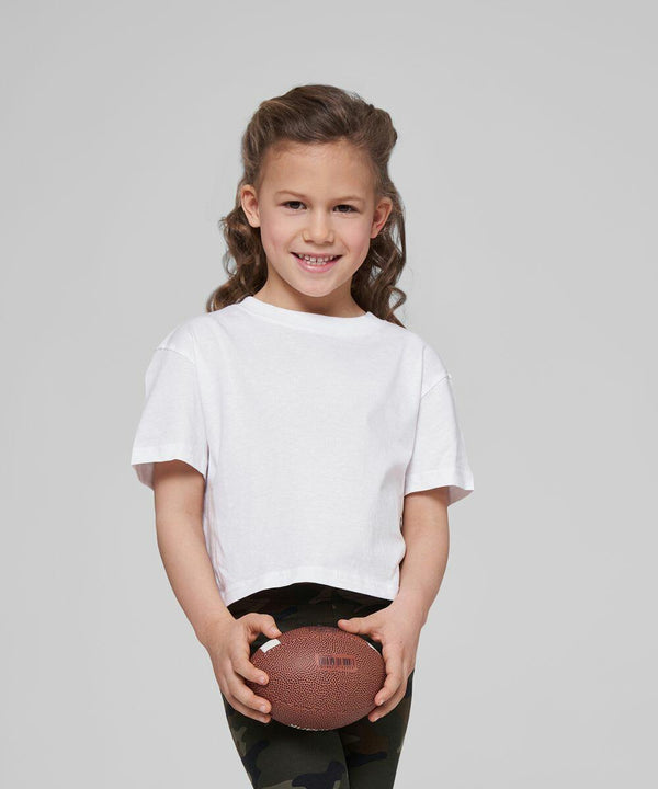 A young girl with curly hair holds a Build Your Brand BY114 in both hands, wearing a white t-shirt and camo-patterned pants, smiling.