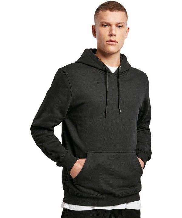 A person is wearing a black Build Your Brand BY118 hooded sweatshirt with drawstrings, standing with hands in the front pockets against a plain white background.
