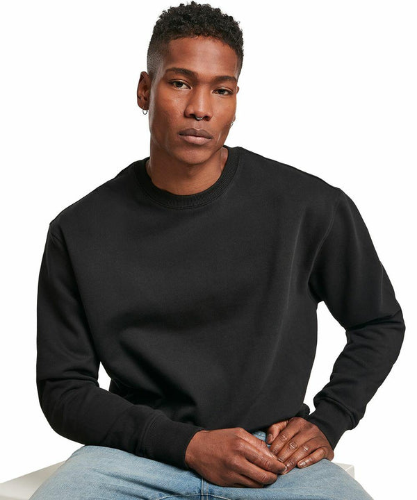 A person is sitting on a white surface, wearing a black Build Your Brand BY120 long-sleeved shirt and blue jeans, looking toward the camera.
