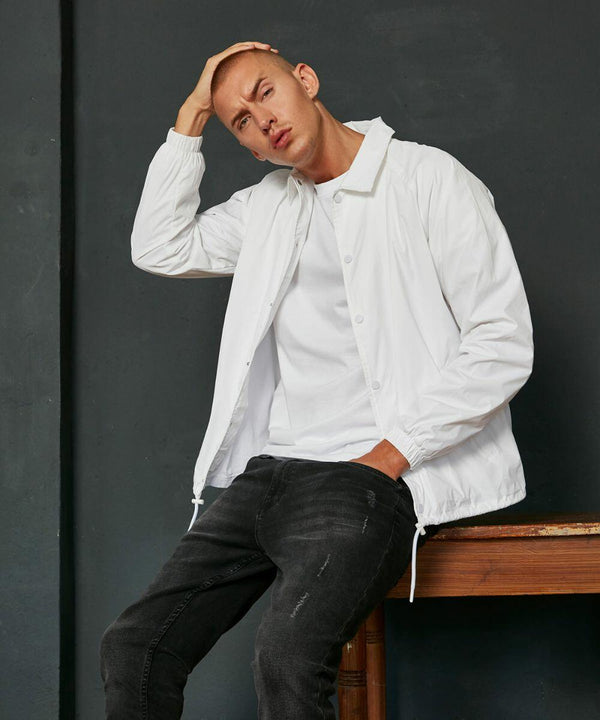 A man in a white jacket and black jeans leans back on a wooden table, with one hand on his head, wearing the BY128 by Build Your Brand.