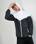 A person wearing a black and white Build Your Brand BY129 hooded jacket stands against a plain wall with one hand on their head.