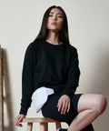 A person with long dark hair sits on a stool, wearing a black sweatshirt and shorts, with a white BY131 accessory from Build Your Brand draped over their leg.