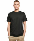 A person stands facing forward with hands in pockets, wearing a Build Your Brand BY136 plain black t-shirt and beige pants against a white background.