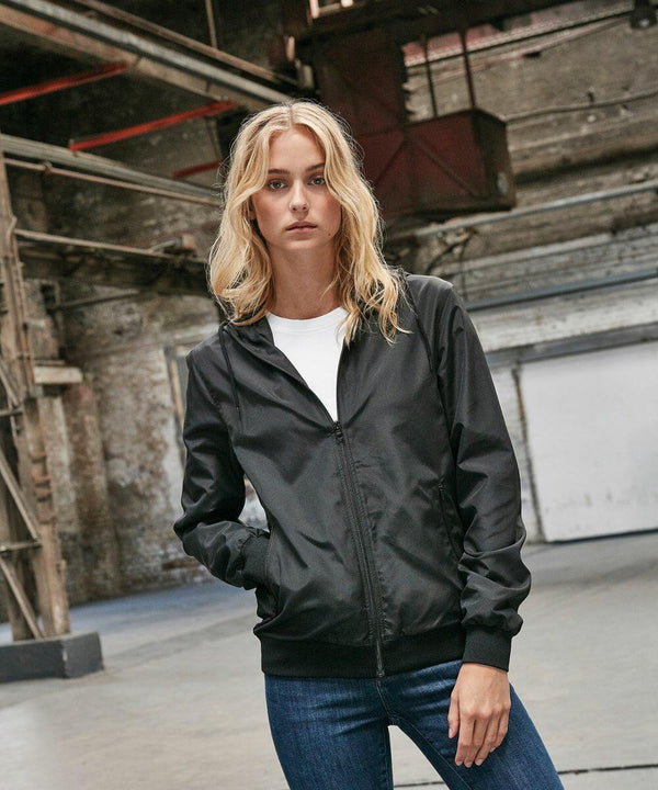 A person with long blond hair, wearing a black jacket over a white shirt and blue jeans, stands in an industrial setting with large metal structures in the background, showcasing the BY147 from Build Your Brand.