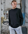 Young man with a buzzcut wearing a Build Your Brand BY150 black long-sleeve shirt and grey sweatpants stands against a graffiti-covered brick wall and industrial background.