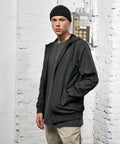 A person dressed in a black beanie, a black Build Your Brand BY151 jacket, a grey shirt, and khaki pants stands indoors against a white brick wall background.