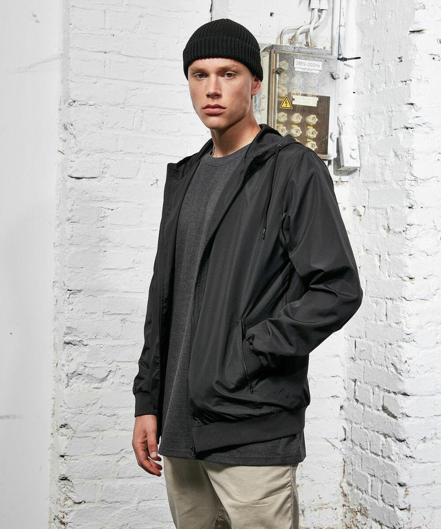 A person dressed in a black beanie, a black Build Your Brand BY151 jacket, a grey shirt, and khaki pants stands indoors against a white brick wall background.
