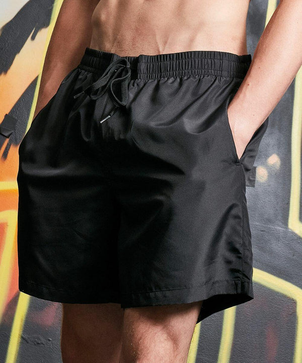 A person wearing Build Your Brand BY153 black drawstring shorts stands with hands in pockets in front of a graffiti-covered wall.