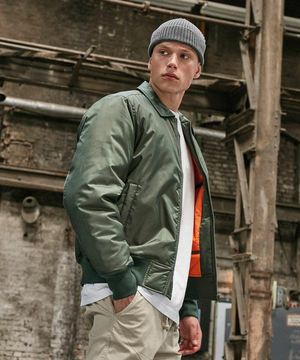A person stands in an industrial setting, wearing a gray beanie, green Build Your Brand BY157 bomber jacket with an orange lining, white shirt, and khaki pants.