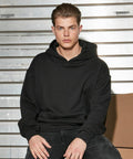 A man wearing a black BY162 hoodie and black pants from Build Your Brand sits in front of a metallic background, looking towards the camera with a neutral expression.