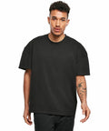 A young man with short curly hair wearing a plain black oversized BY163 T-shirt by Build Your Brand and black pants stands facing the camera. He has a tattoo on his left forearm.