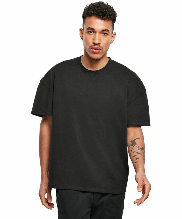 A young man with short curly hair wearing a plain black oversized BY163 T-shirt by Build Your Brand and black pants stands facing the camera. He has a tattoo on his left forearm.