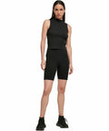 A person in a black sleeveless top, black BY184 biker shorts by Build Your Brand, and black sneakers stands on a plain white background.