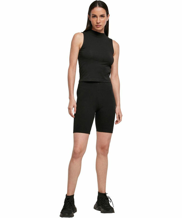 A person in a black sleeveless top, black BY184 biker shorts by Build Your Brand, and black sneakers stands on a plain white background.