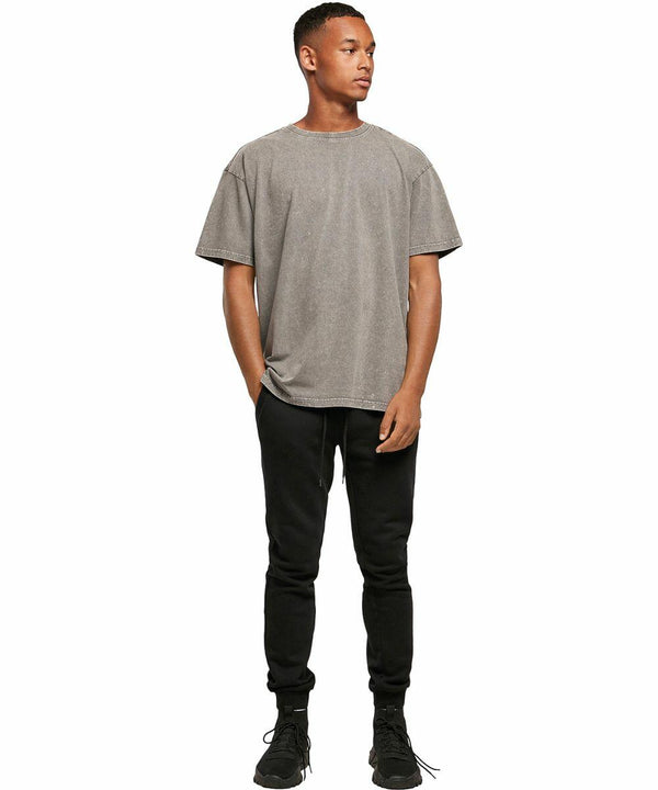A man stands wearing a grey BY189 T-shirt from Build Your Brand and black pants, with black shoes, posing against a plain white background.
