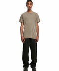A person wearing a plain brown BY190 T-shirt by Build Your Brand, black cargo pants, and black shoes stands against a white background.