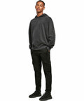 A person stands with hands in pockets, wearing a dark gray hoodie, black cargo pants, and black Build Your Brand BY191 sneakers, against a plain white backdrop.