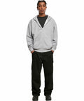 A person stands facing forward wearing a grey BY192 hoodie by Build Your Brand, black t-shirt, black pants, and black shoes, with hands in pockets, against a white background.