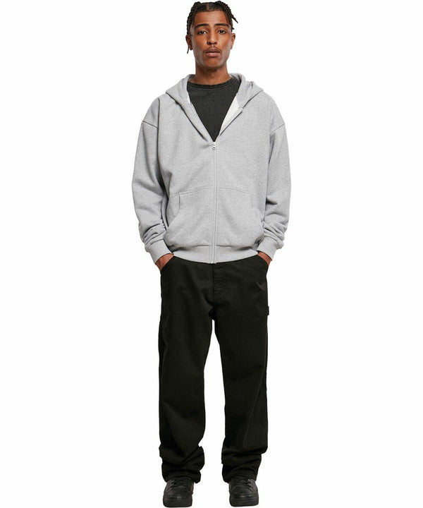 A person stands facing forward wearing a grey BY192 hoodie by Build Your Brand, black t-shirt, black pants, and black shoes, with hands in pockets, against a white background.