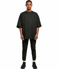 A person is standing wearing a black oversized t-shirt by Build Your Brand, model BY193, black pants, white socks, and black sneakers, posing against a plain white background.