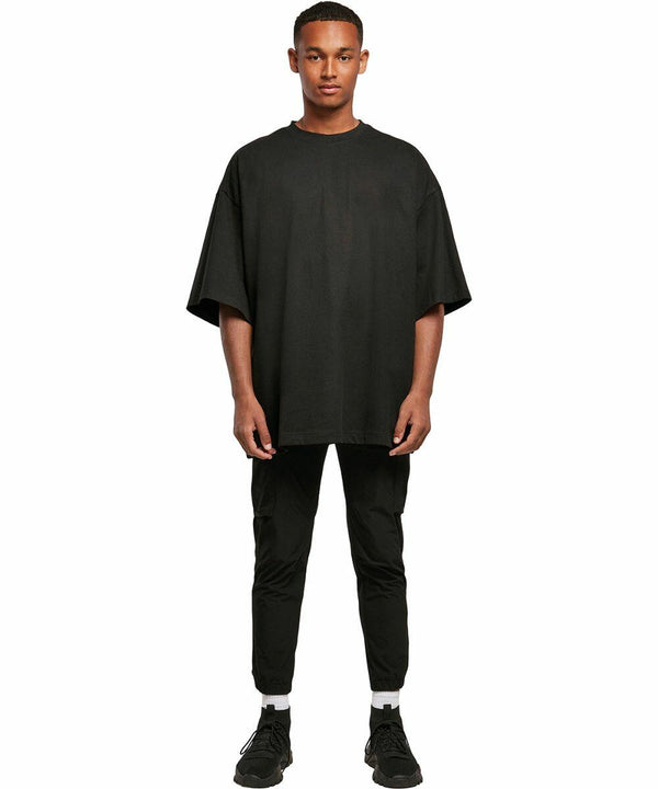 A person is standing wearing a black oversized t-shirt by Build Your Brand, model BY193, black pants, white socks, and black sneakers, posing against a plain white background.