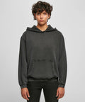 Person with short curly hair wearing a dark gray Build Your Brand BY194 hoodie with a front pocket, standing against a plain background.
