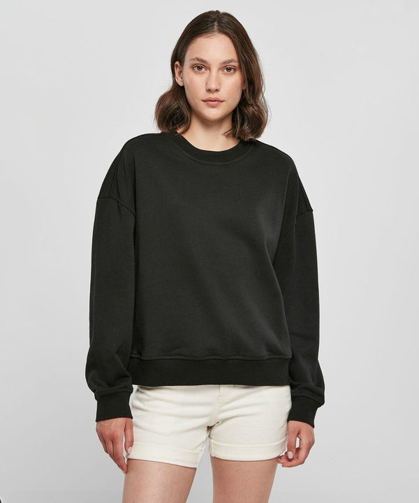 A person with shoulder-length hair is wearing a black Build Your Brand BY212 sweatshirt and white shorts, standing against a plain background.