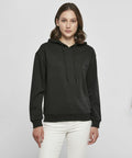A person with shoulder-length hair wearing a black BY213 hoodie by Build Your Brand and white pants stands against a neutral background.