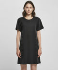 A woman is standing and facing forward, wearing a plain black short-sleeved dress from Build Your Brand, specifically the BY214 model. She has shoulder-length brown hair and a neutral expression. The background is plain white.