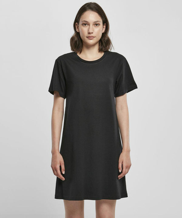 A woman is standing and facing forward, wearing a plain black short-sleeved dress from Build Your Brand, specifically the BY214 model. She has shoulder-length brown hair and a neutral expression. The background is plain white.