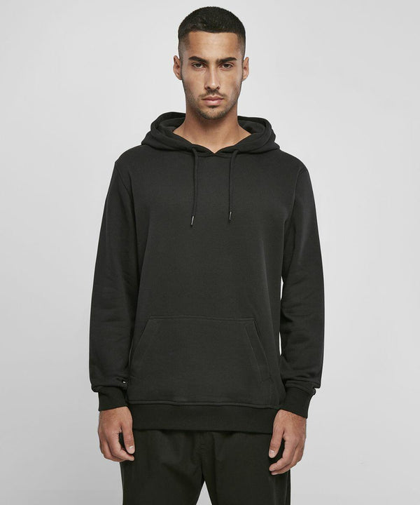 A man stands facing forward, wearing a Build Your Brand BY215 hoodie with a front pouch pocket. The background is plain and light-colored.