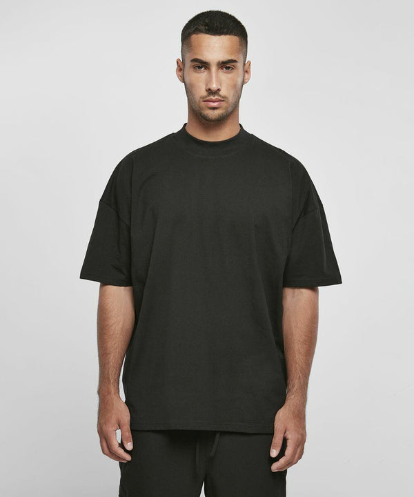 A man stands facing the camera, wearing a Build Your Brand BY230 and black pants against a neutral background.
