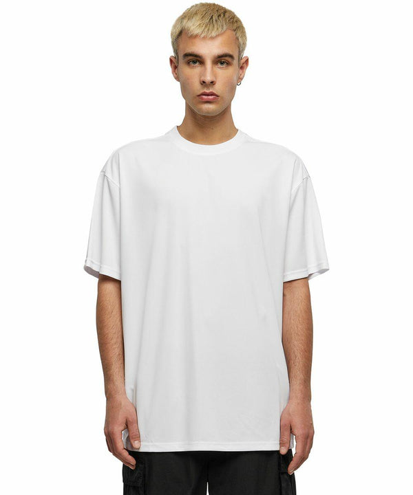 A person with short, light blond hair wears a plain white oversized Build Your Brand BY249 T-shirt and black pants, standing against a white background.