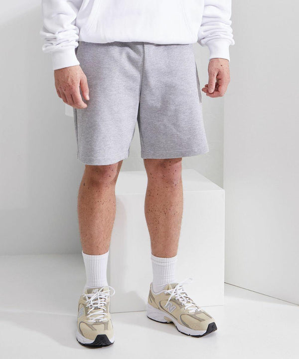 A person wearing a white BY251 hoodie from Build Your Brand, light gray shorts, white socks, and beige New Balance sneakers standing indoors.