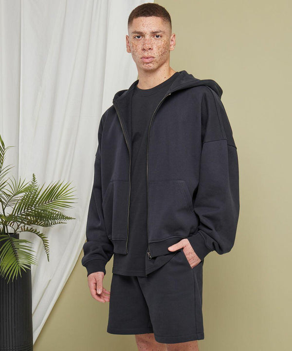 Person standing indoors, wearing a black BY254 hoodie with the hood down, black t-shirt, and black shorts by Build Your Brand. One hand is in their pocket. There is a potted plant and a white curtain in the background.