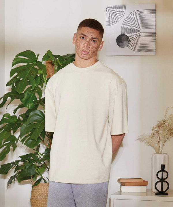 A person stands indoors wearing a beige BY256 t-shirt by Build Your Brand and grey pants, next to a potted plant and a small table. A minimalist artwork is mounted on the wall behind them.