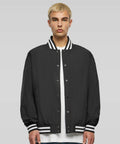 A person with short blond hair wearing a black BY259 varsity jacket by Build Your Brand over a white shirt stands against a plain background.