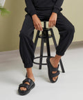 Person sitting on a black stool, wearing black jogger pants and BY265 slide sandals by Build Your Brand, with hands resting on thighs.