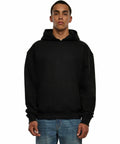 A person wearing a Build Your Brand BY268 plain black hoodie and blue jeans standing against a white background.