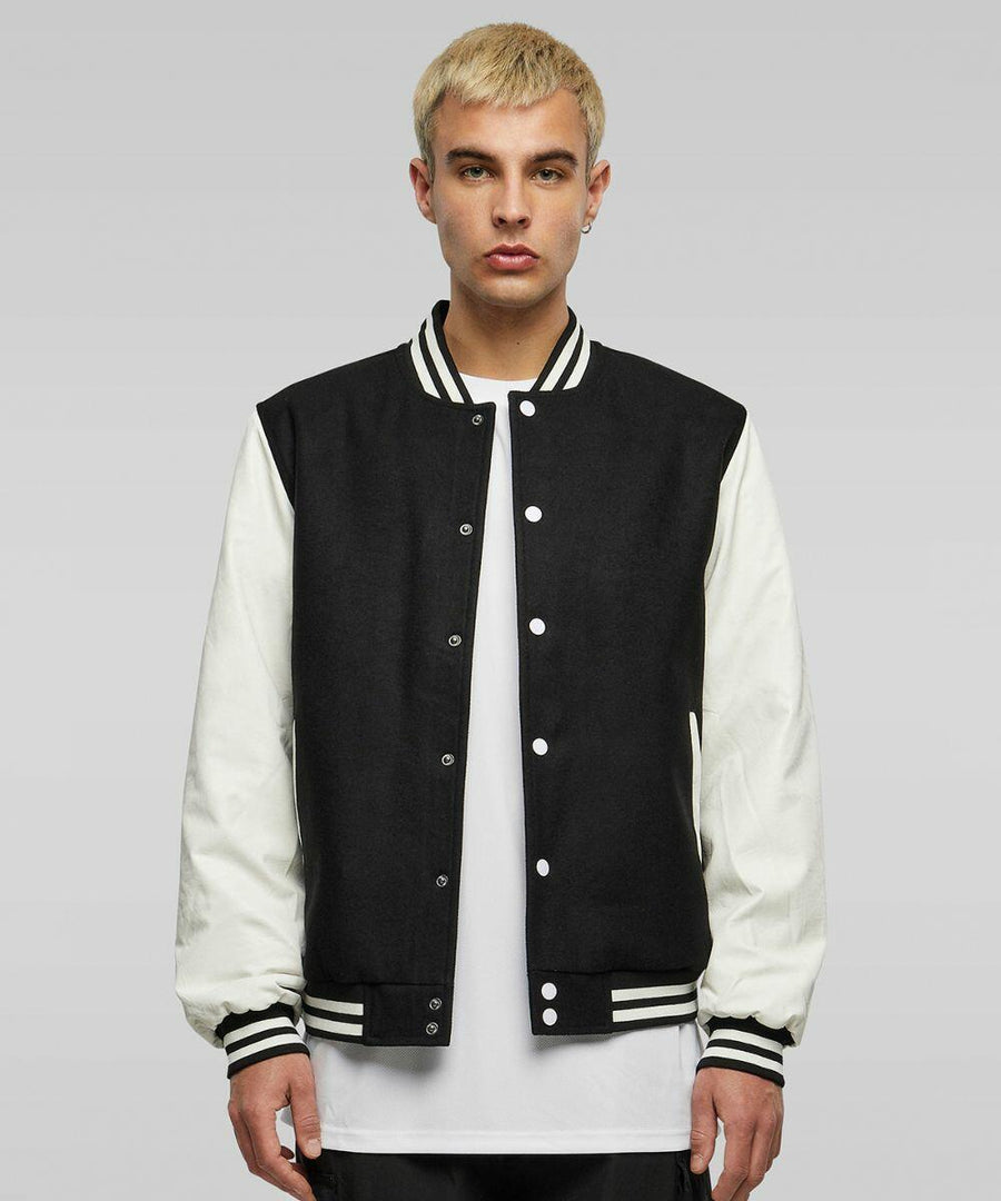 A person with short blonde hair is standing and wearing a black varsity jacket with white sleeves over a white T-shirt from Build Your Brand, product model BY269. The background is plain gray.