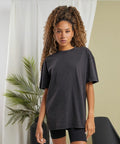 A person with long, curly hair stands in front of a white curtain and a palm plant, wearing a loose, dark grey BY270 t-shirt and black shorts by Build Your Brand.