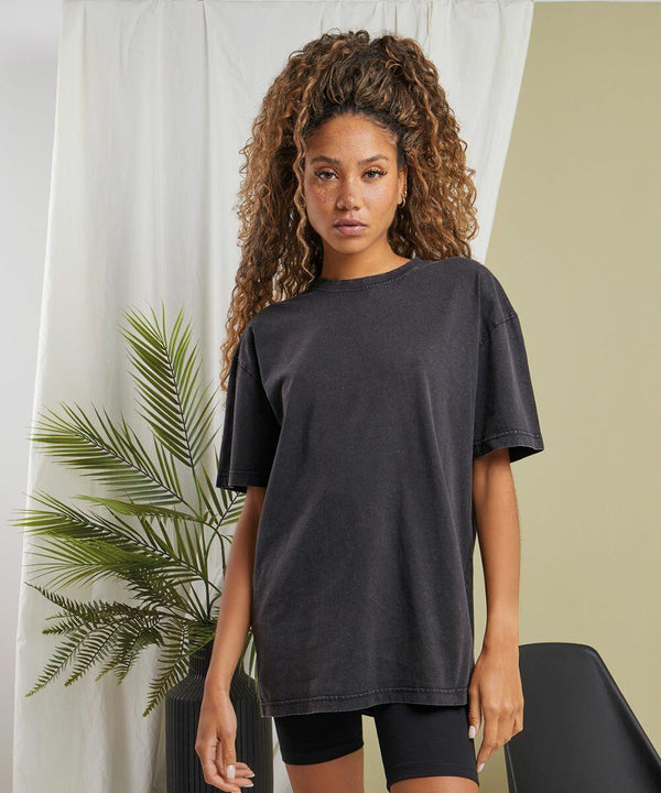A person with long, curly hair stands in front of a white curtain and a palm plant, wearing a loose, dark grey BY270 t-shirt and black shorts by Build Your Brand.