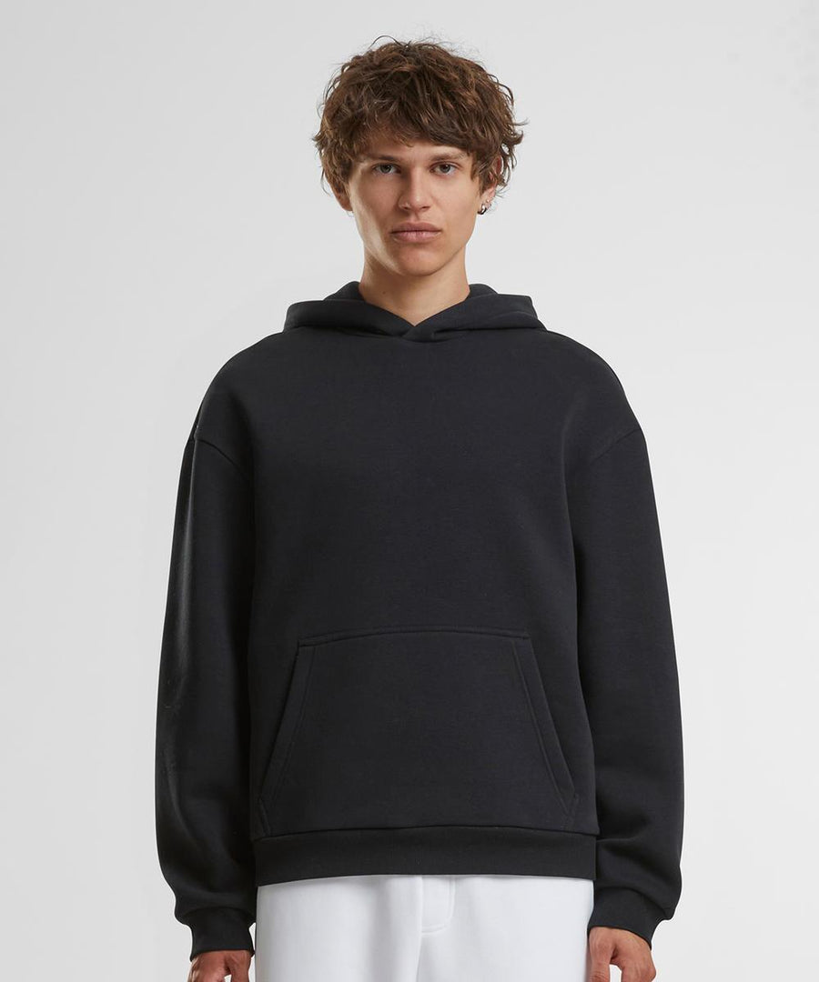 A person with short, curly brown hair wears the Build Your Brand Fluffy hoodie in black and white trousers, standing against a plain white background and facing the camera.
