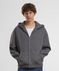 A person with short brown hair wears the Build Your Brand Fluffy zip hoodie in grey over a white shirt, standing against a plain white background with hands in the hoodie pockets.