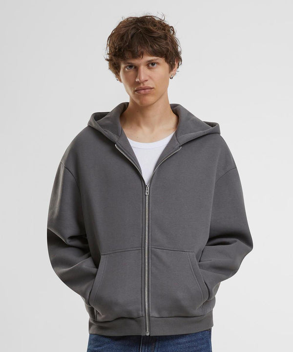 A person with short brown hair wears the Build Your Brand Fluffy zip hoodie in grey over a white shirt, standing against a plain white background with hands in the hoodie pockets.