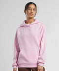 A woman with her hair tied back stands against a plain background wearing the Build Your Brand Women's Fluffy Hoodie in light pink, featuring a hood and front pocket, paired with brown trousers as she gazes slightly to the side.