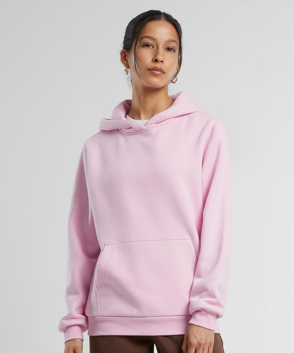 A woman with her hair tied back stands against a plain background wearing the Build Your Brand Women's Fluffy Hoodie in light pink, featuring a hood and front pocket, paired with brown trousers as she gazes slightly to the side.