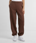 A person wears Build Your Brand Women's fluffy joggers in brown, paired with a white top and white trainers, standing with hands in pockets against a plain light background.
