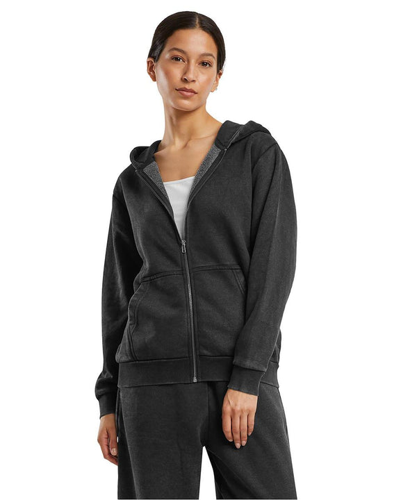 A person wears the Build Your Brand Women's vintage heavy zip hoodie in dark grey with front pockets and a hood, styled over a white top with matching trousers, against a plain white background.